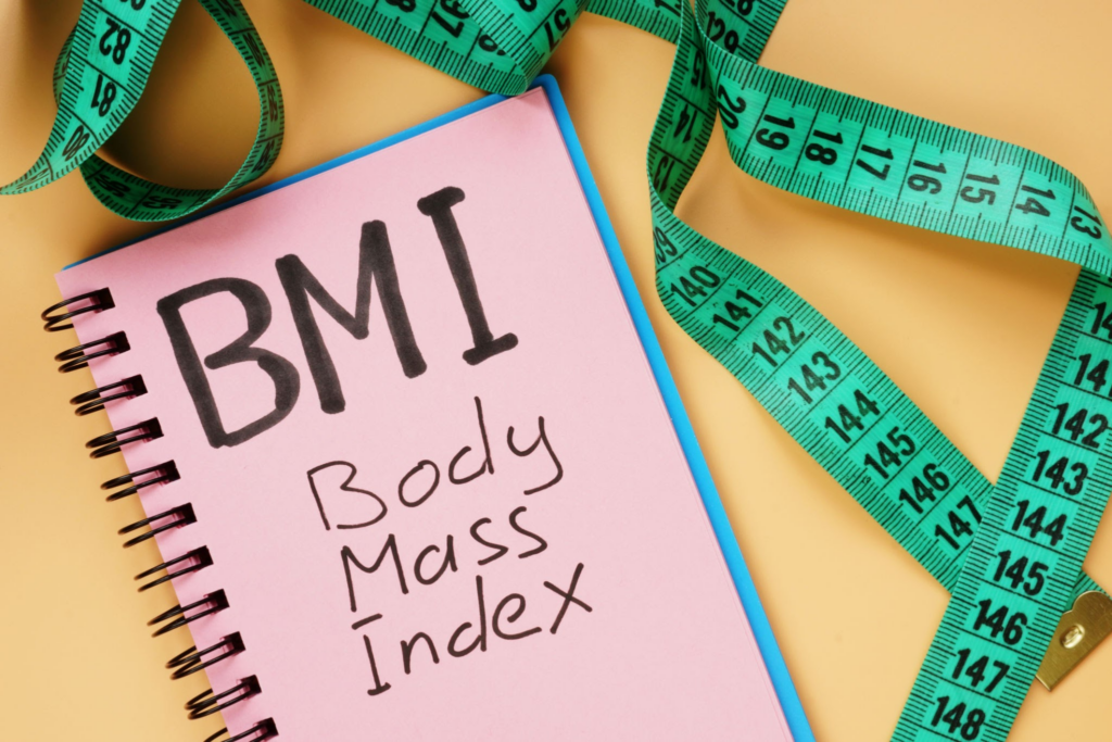 A pink notepad with the text 'BMI Body Mass Index' written on it, placed on a yellow background with a green measuring tape coiled around it.