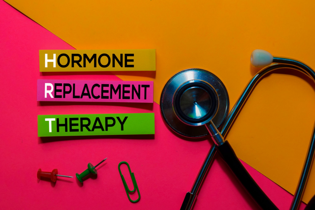 A vibrant image featuring the phrase 'HORMONE REPLACEMENT THERAPY' written on colorful sticky notes in yellow, pink, and green, arranged on a bright pink and orange background. The image includes a stethoscope lying on the right side with its earpieces visible, along with two pushpins (one red and one green) and a green paperclip at the bottom left corner.