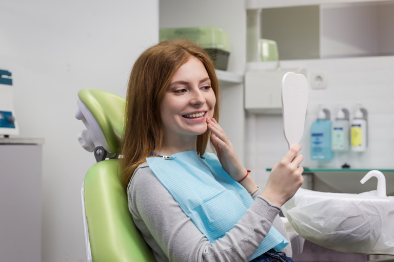 Una mujer sonriente, de cabello rojo, sentada en un sillón dental verde, con un babero dental azul claro, mirándose en un espejo de mano mientras se toca la mejilla.