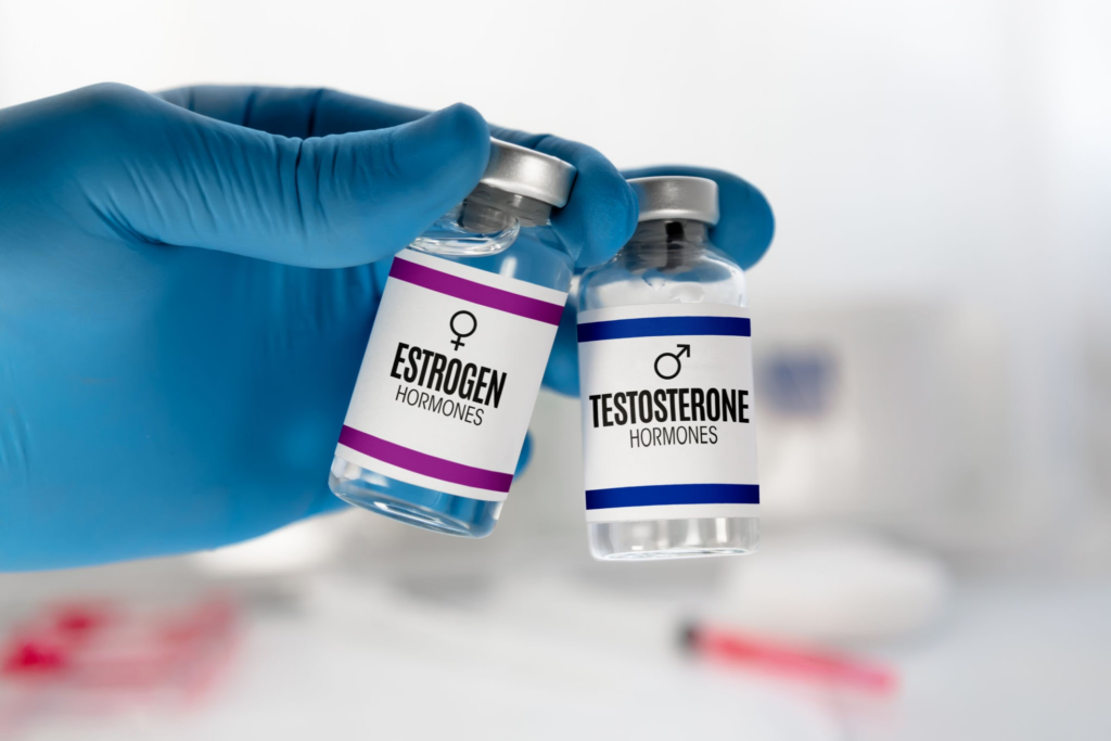 Una mano guantata che tiene due fiale etichettate "Ormoni estrogeni" e "Ormoni testosterone" in un ambiente medico o di laboratorio.