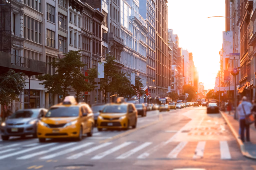 Una bulliciosa calle de la ciudad de Nueva York, con taxis amarillos y peatones, flanqueada por altos edificios y árboles, capturada por la cálida luz del atardecer.