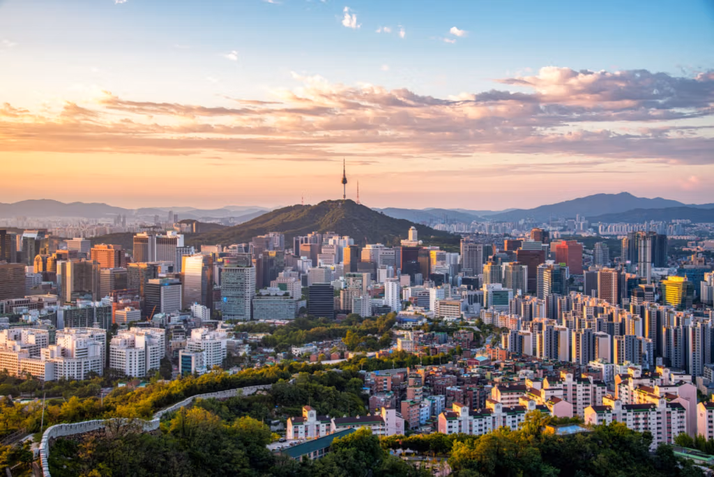 Vista panorámica de Seúl, Corea del Sur, al atardecer, con la Torre N de Seúl destacando en una colina al fondo. El paisaje urbano presenta una mezcla de modernos rascacielos y edificios residenciales, con montañas visibles a lo lejos. La escena está bañada por una cálida luz dorada, creando un paisaje urbano sereno y pintoresco.