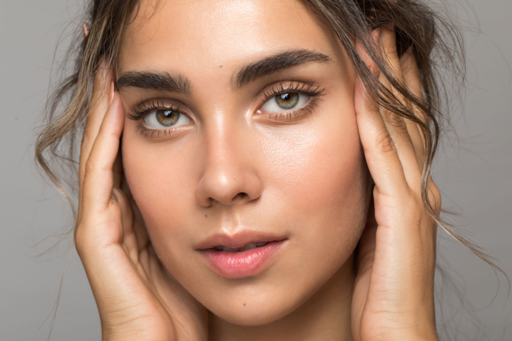 Nahaufnahme einer jungen Frau mit reiner Haut, die ihr Gesicht mit beiden Händen hält. Sie hat grüne Augen, dichte Augenbrauen und welliges braunes Haar. Der Hintergrund ist schlicht und hell und lenkt den Blick auf ihre Gesichtszüge.