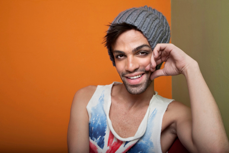Un hombre sonriente que viste un gorro gris y una camiseta sin mangas con un diseño de bandera posa sobre un fondo naranja.