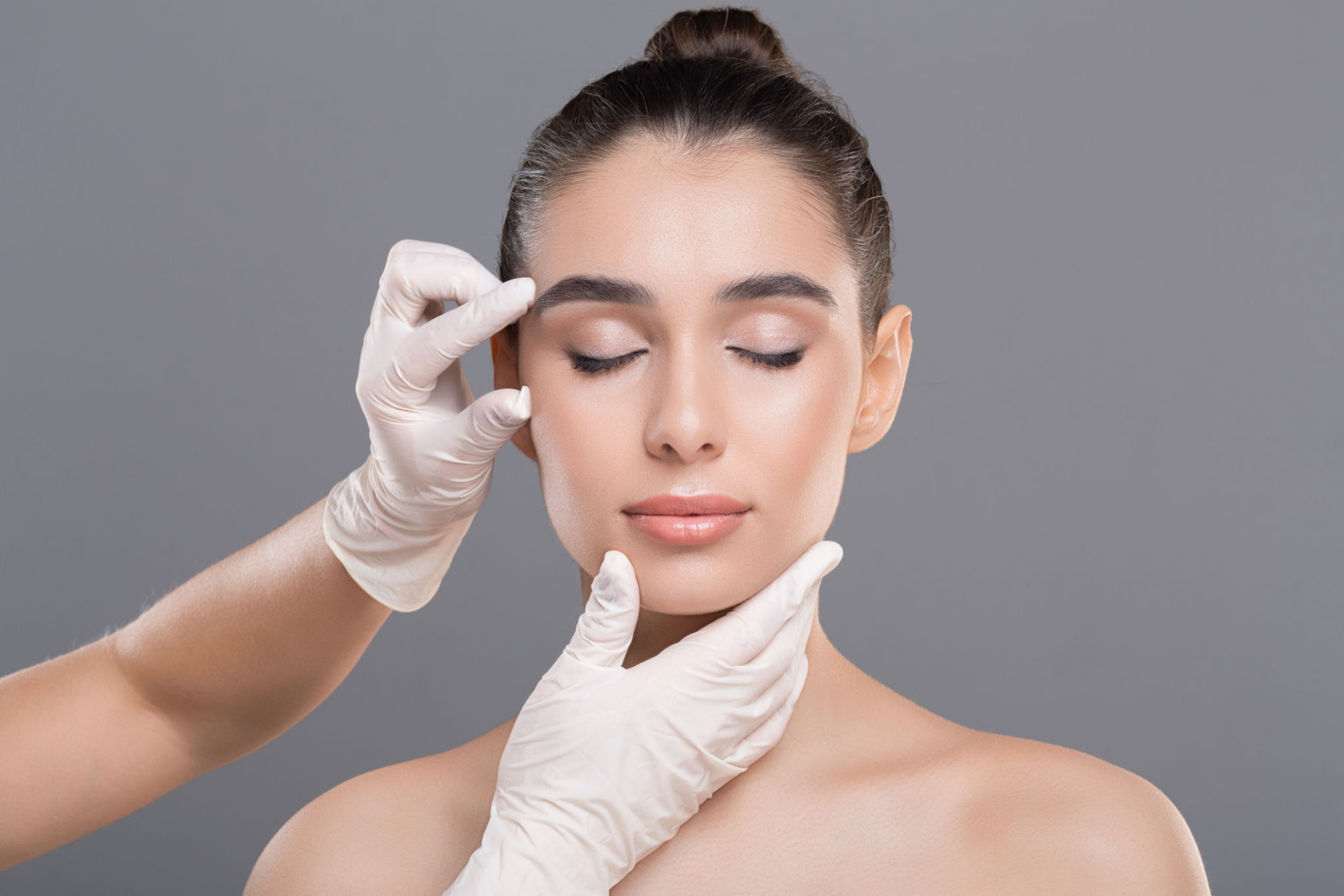 Una donna che si sottopone a un esame o trattamento del viso da parte di un professionista che indossa guanti, mentre le mani del professionista le toccano delicatamente la guancia e la fronte.