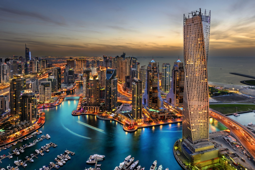 Eine atemberaubende Luftaufnahme der Dubai Marina bei Sonnenuntergang. Der ikonische Cayan Tower und die umliegenden Wolkenkratzer spiegeln sich im ruhigen Wasser, entlang der Marina liegen Boote, und die Lichter der Stadt beginnen, die Szene zu erhellen.