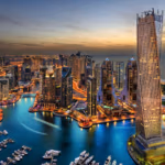 A stunning aerial view of Dubai Marina at sunset, showcasing the iconic Cayan Tower and surrounding skyscrapers reflected in the tranquil waters, with boats docked along the marina and the city's lights beginning to illuminate the scene.