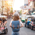 Tayland'ın başkenti Bangkok'ta, renkli tuk-tuk'lar ve şehir binalarıyla çevrili, hareketli bir sokakta şapka ve sırt çantası takmış bir kadın duruyor.