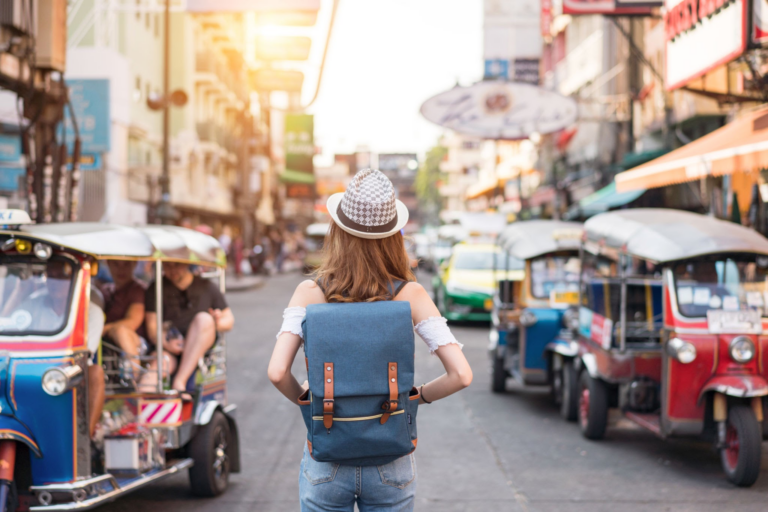 Una mujer con sombrero y mochila se encuentra en una bulliciosa calle de Bangkok, Tailandia, rodeada de coloridos tuk-tuks y edificios de la ciudad.