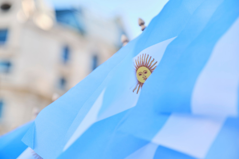Primer plano de una bandera azul con un sol amarillo en el centro, sobre un fondo urbano borroso. La bandera parece ondear ligeramente, y el sol tiene un rostro humanoide con rayos que se extienden hacia afuera.