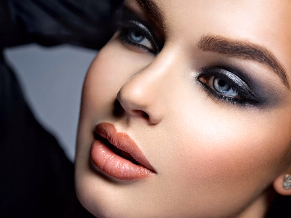 A close-up portrait of a woman with striking blue eyes, wearing dramatic smoky eye makeup, nude lipstick, and a black headscarf. The image focuses on her facial features, highlighting her makeup and expression.