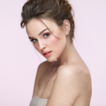 Een vrouw met elegant opgestoken haar, subtiele make-up en een vriendelijke blik over haar schouder tegen een effen achtergrond.