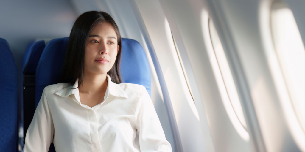 Una mujer sentada en un asiento de avión azul, mirando por la ventana con una expresión pensativa, vistiendo una camisa blanca.
