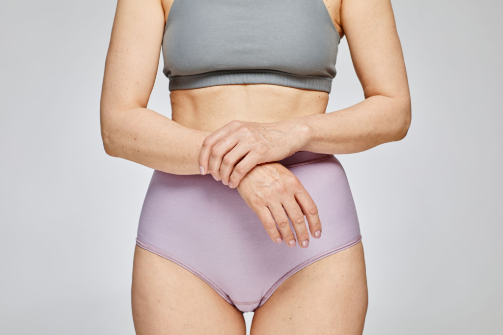 Une personne portant un soutien-gorge de sport gris et un slip taille haute lavande, les mains posées sur les hanches.