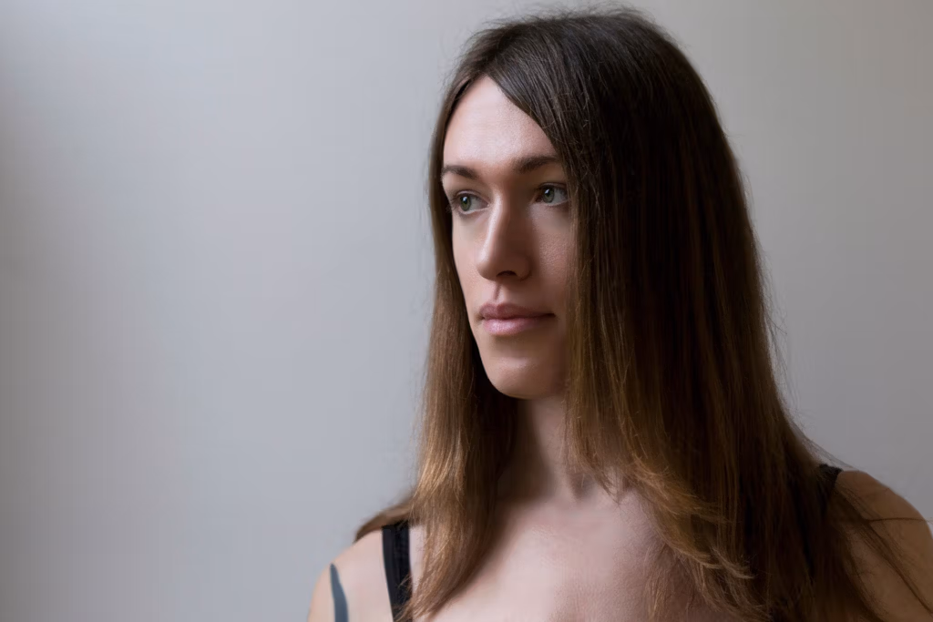 Eine junge Frau mit langen braunen Haaren blickt direkt in die Kamera. Sie trägt ein ärmelloses Oberteil vor einem schlichten hellen Hintergrund.