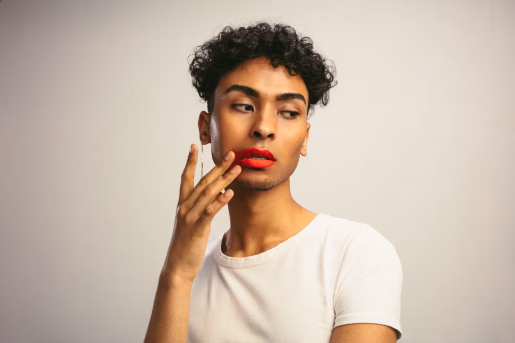 A person with curly hair and red lipstick wearing a white t-shirt, posing thoughtfully with their hand on their face.