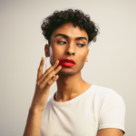 Eine Person mit lockigem Haar und rotem Lippenstift, die ein weißes T-Shirt trägt und nachdenklich mit der Hand vor dem Gesicht posiert.