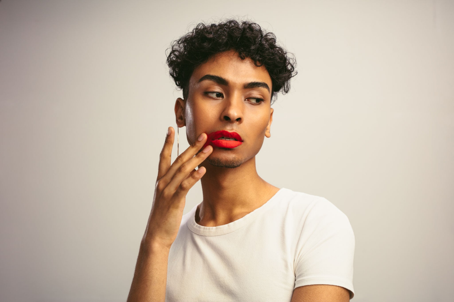 A person with curly hair and red lipstick wearing a white t-shirt, posing thoughtfully with their hand on their face.