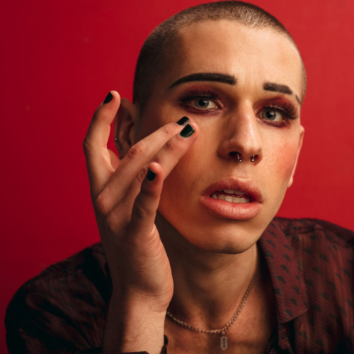A person with a shaved head and dramatic eye makeup, wearing a dark patterned top, poses against a red background while gently touching their face with black-painted nails.