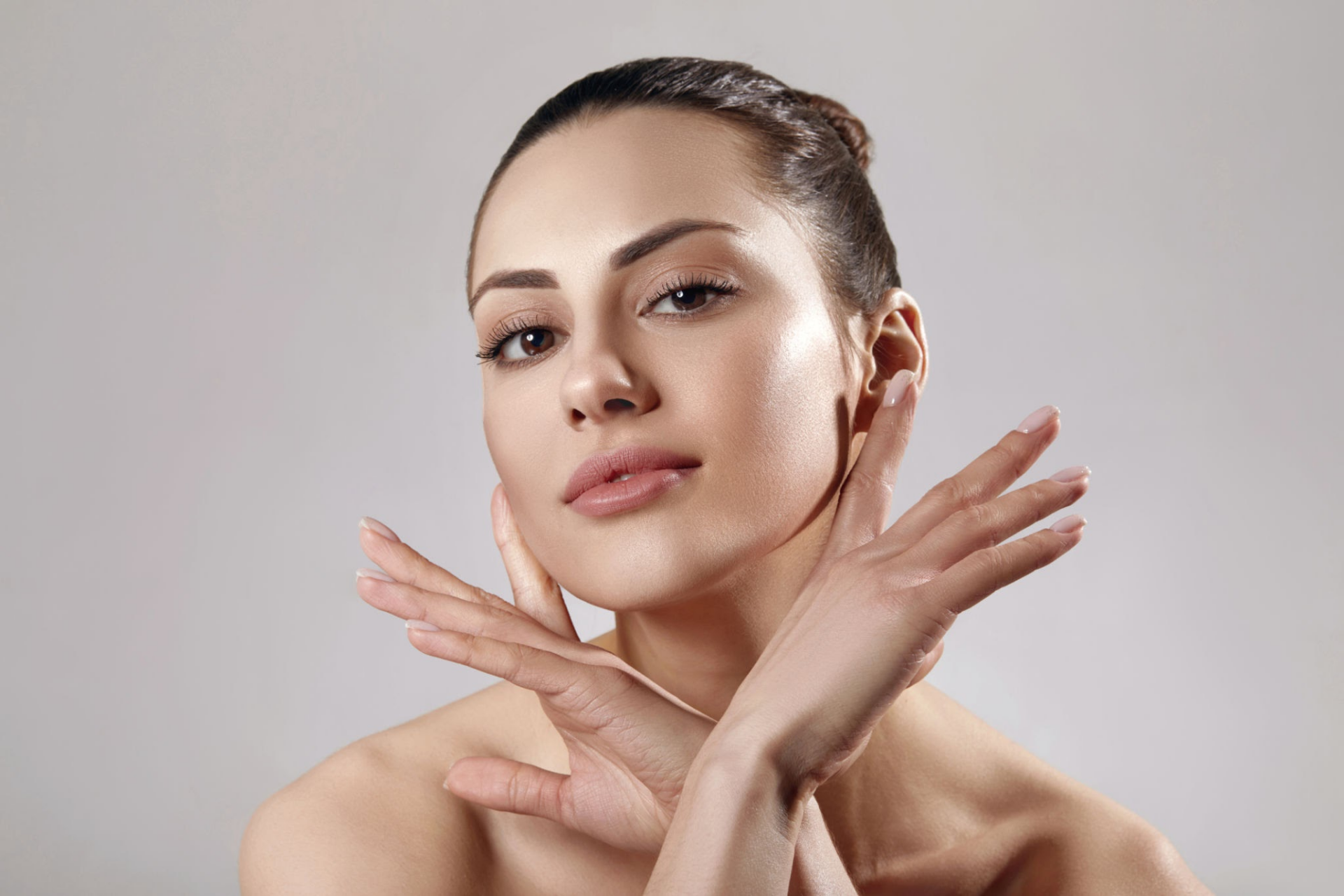 A woman with her hands gently touching her face, showcasing smooth skin and a serene expression, against a neutral background.