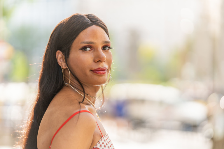 Una mujer joven con cabello largo y oscuro y aretes de aro que luce un top de lunares rojos y blancos parada al aire libre en un entorno natural iluminado por el sol.