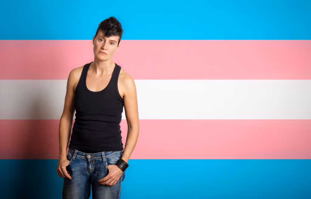 Eine Person steht selbstbewusst vor einem Hintergrund mit der Transgender-Pride-Flagge, die aus horizontalen Streifen in Blau, Rosa und Weiß besteht.