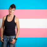 Een persoon staat zelfverzekerd voor een achtergrond met de transgender pride-vlag, die bestaat uit horizontale strepen in blauw, roze en wit.