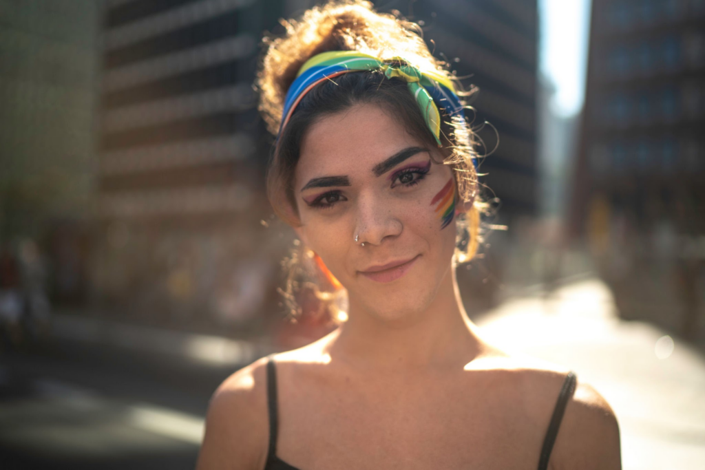 Eine junge Frau mit lockigem Haar, das mit einem bunten Stirnband zurückgebunden ist, trägt ein schwarzes Tanktop und hat Regenbogen-Gesichtsbemalung auf der Wange. Sie lächelt in einer Umgebung im Freien.