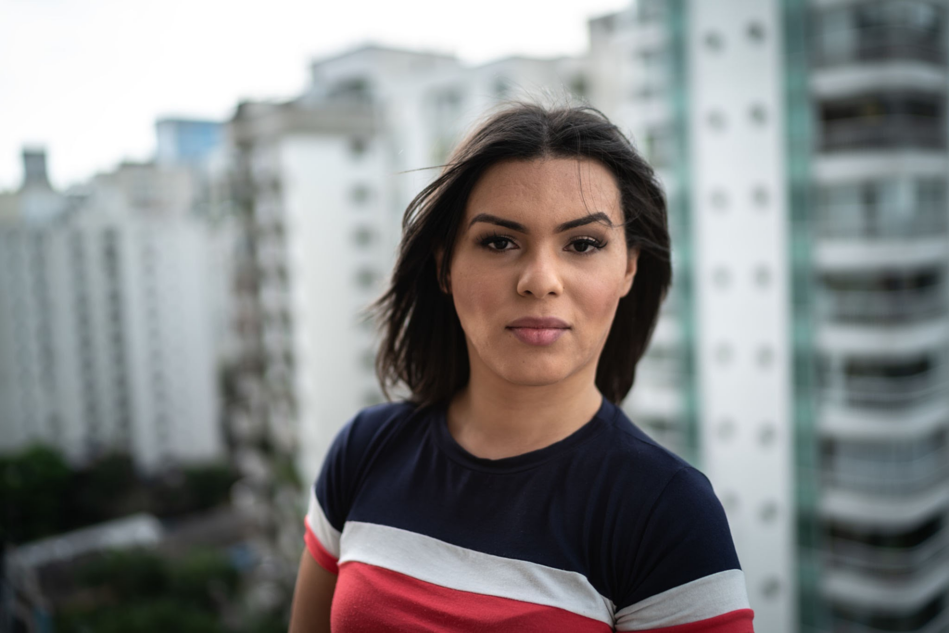 Kısa saçlı, çizgili gömlek giymiş genç bir kadın, arka planda yüksek binaların bulunduğu kentsel bir ortamda dışarıda duruyor.