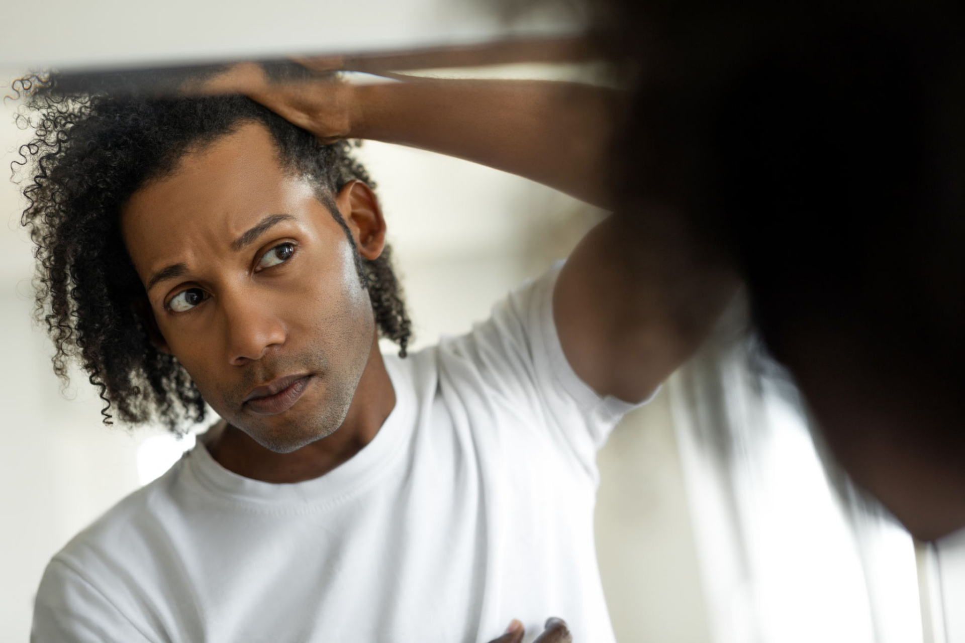 Een man met krullend haar bekijkt zichzelf in de spiegel en schikt met één hand zijn haar.