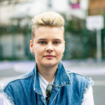 Une personne aux cheveux blonds courts portant une veste en jean sur une chemise blanche, debout dans une rue de la ville avec des bâtiments flous en arrière-plan.