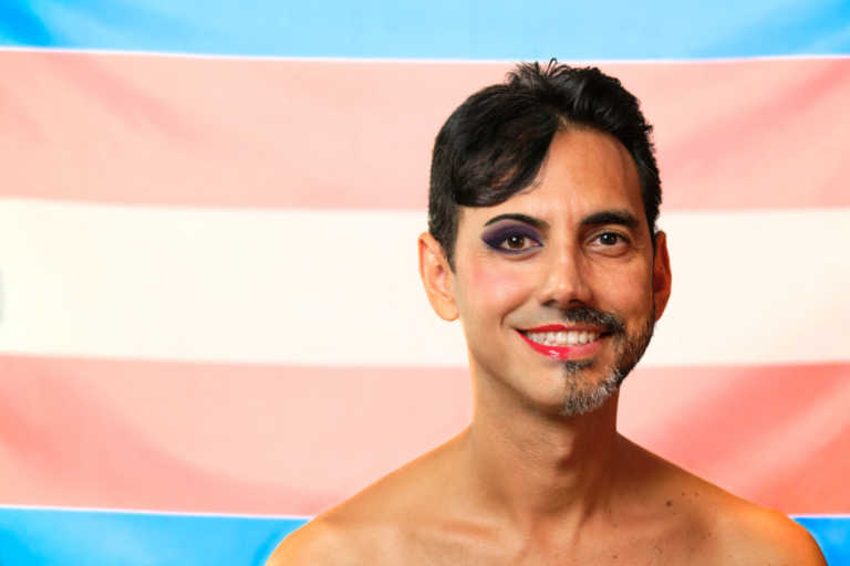 Una persona con cabello corto y oscuro, vello facial y maquillaje, que incluye sombra de ojos violeta y lápiz labial rojo, sonriendo frente a un fondo con rayas horizontales en rosa, blanco y azul, que representan la bandera del orgullo transgénero.