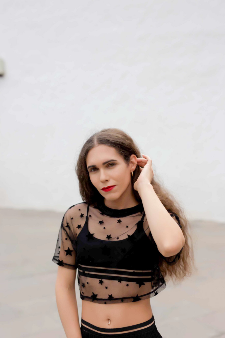 Женщина с красной помадой на губах, одетая в черный сетчатый топ со звездным рисунком, позирует на улице в яркий день.