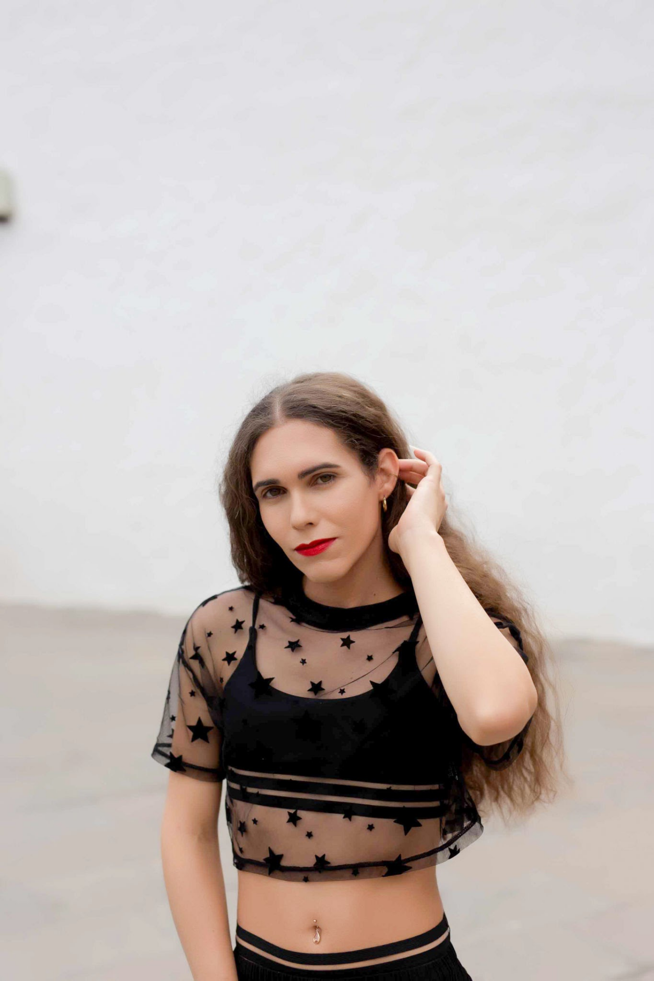 A woman with red lipstick wearing a black mesh top with star patterns, posing outdoors on a bright day.