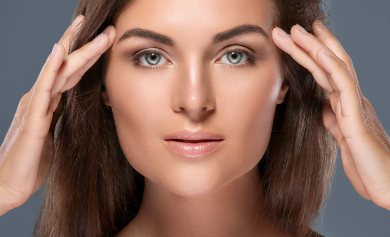 Una fotografía de primer plano de una mujer con una piel suave y radiante, sujetándose el cabello hacia atrás con ambas manos mientras mira directamente a la cámara.