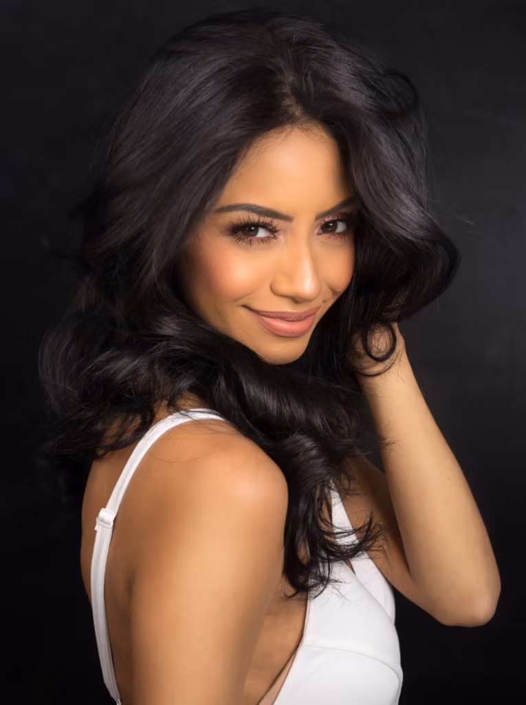 A woman with long, dark curly hair wearing a white dress posing against a dark background.