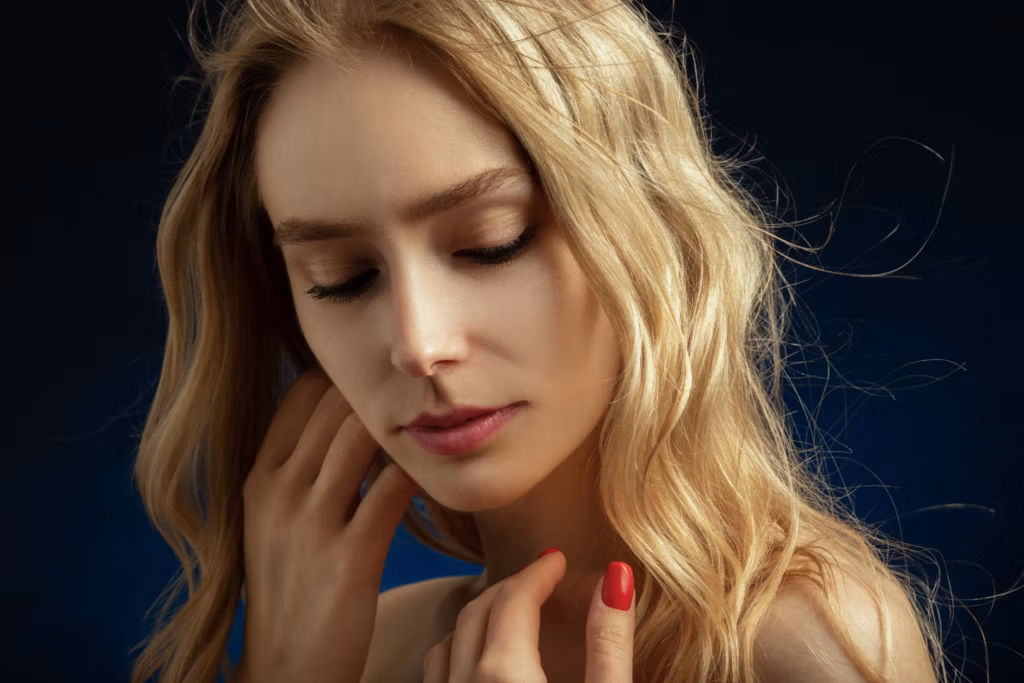Ein Nahaufnahmeporträt einer Frau mit langen blonden Haaren und rotem Nagellack, die vor einem dunklen Hintergrund nachdenklich wirkt.