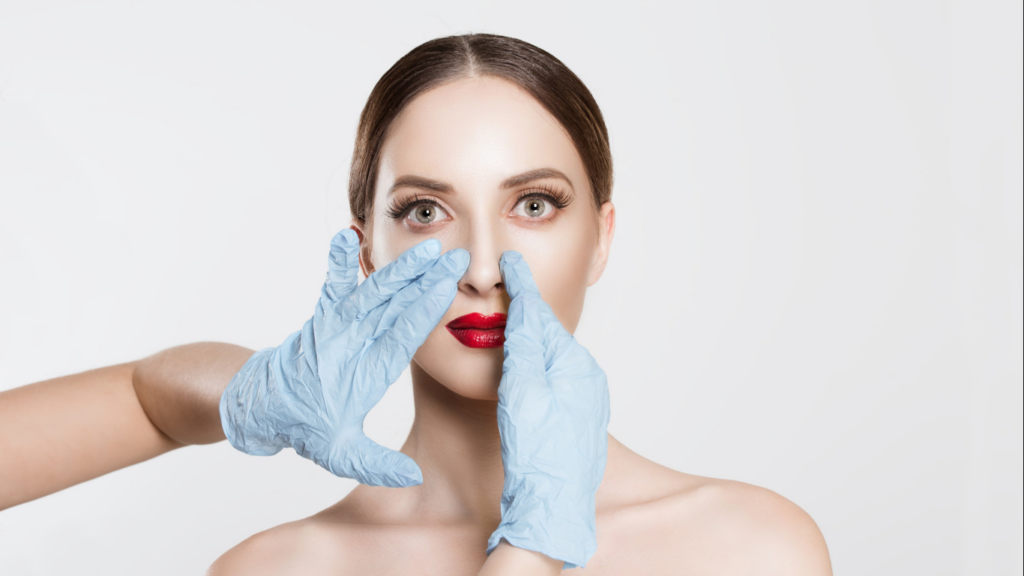 On voit une femme avec du rouge à lèvres rouge se tenir le nez à deux mains, portant des gants médicaux bleus.