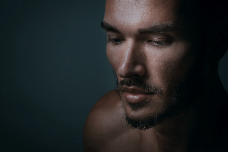 Retrato en primer plano de un hombre con barba y cabello corto, mirando ligeramente hacia abajo con expresión seria. La iluminación proyecta un lado de su rostro en sombra, resaltando la textura de su piel y sus rasgos faciales. El fondo es oscuro, resaltando su rostro.