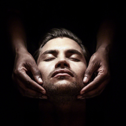 A man receiving a relaxing facial massage, with hands gently placed on his face, against a dark background.