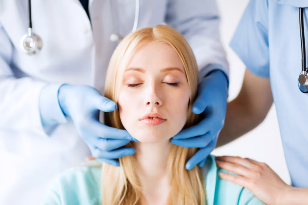 Two medical professionals in scrubs and gloves carefully examining a patient's face, likely in a clinical setting.