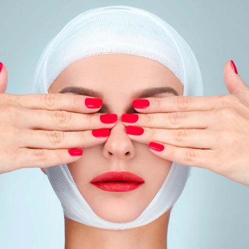 A person with their face partially covered by their hands, wearing a white head wrap and red nail polish, against a light blue background.
