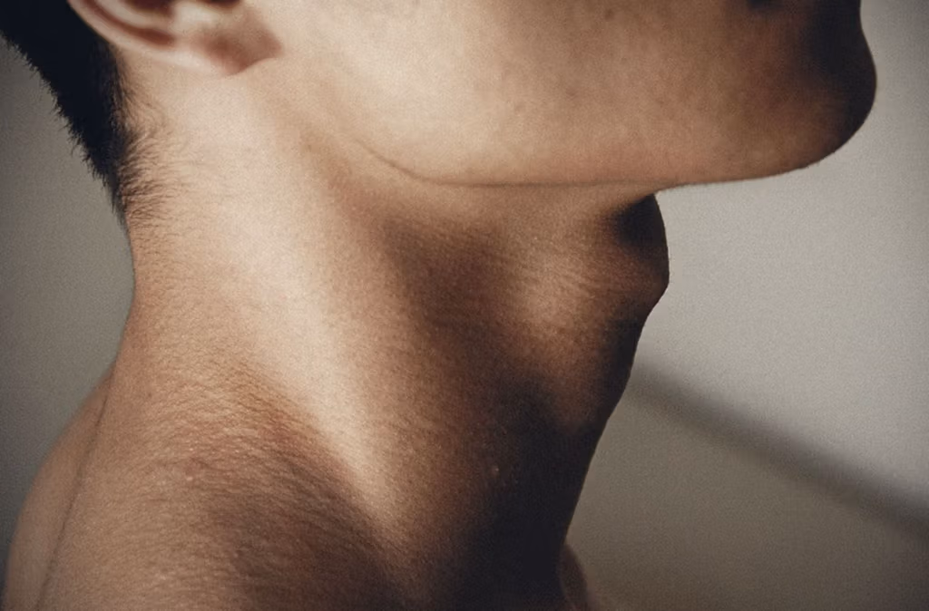 Primer plano del cuello y la mandíbula inferior de una persona con vello facial corto, resaltando la textura de la piel sobre un fondo liso.