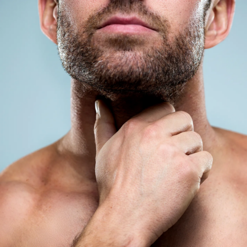 A close-up image of a bearded man touching his neck, possibly examining his thyroid area or Adam's apple. The man has short facial hair and a focused expression, with his fingers gently placed on his throat. The background is plain and neutral, emphasizing the subject's action and facial features.