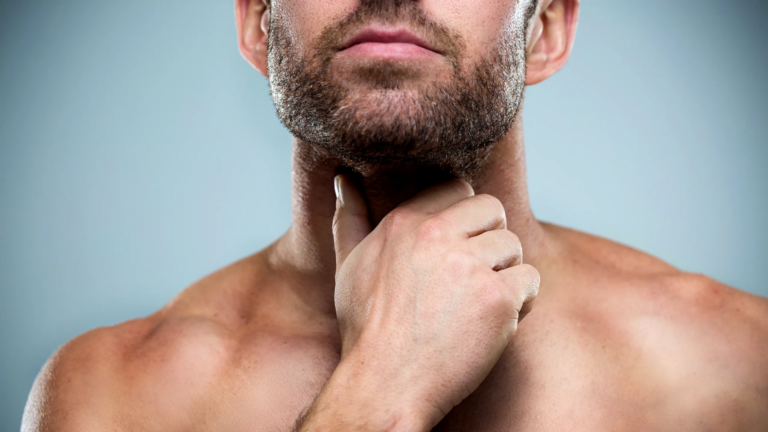 Primer plano de un hombre con barba tocándose el cuello, posiblemente examinando la tiroides o la nuez de Adán. El hombre tiene vello facial corto y una expresión concentrada, con los dedos suavemente apoyados en la garganta. El fondo es liso y neutro, lo que enfatiza la acción y los rasgos faciales del sujeto.