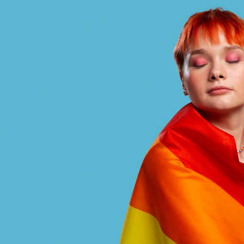 A person with short orange hair and pink eyeshadow, draped in a vibrant rainbow flag, stands against a solid blue background, symbolizing pride and individuality.
