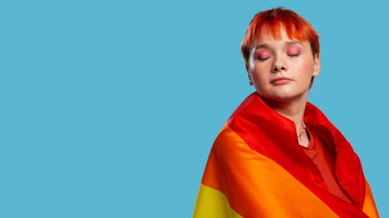 Una persona con cabello corto de color naranja y sombra de ojos rosa, envuelta en una vibrante bandera de arcoíris, se encuentra sobre un fondo azul sólido, que simboliza el orgullo y la individualidad.