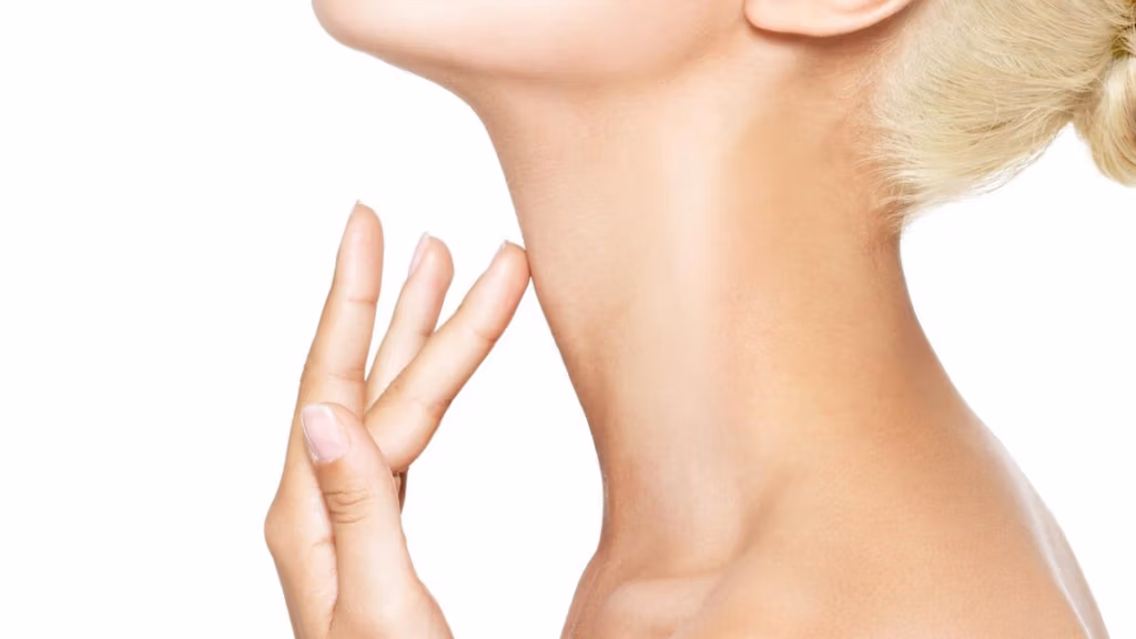 Primer plano del cuello y la mano de una mujer. La mujer se toca suavemente el cuello con las yemas de los dedos, posiblemente buscando signos de envejecimiento o alguna condición de la piel. La atención se centra en la suavidad y textura de la piel, destacando la zona del cuello.
