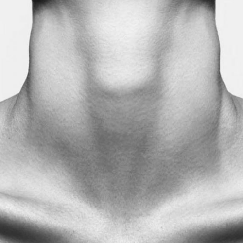 Close-up black and white image of a person's neck and collarbone area, showing smooth skin with a small mole on the left shoulder. The image is cropped to focus on the neck and upper chest, with a plain white background.