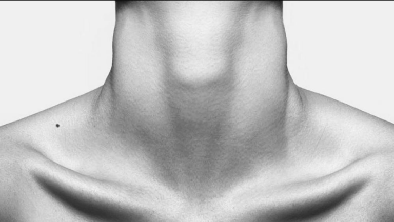 Primer plano en blanco y negro del cuello y la clavícula de una persona, con piel lisa y un pequeño lunar en el hombro izquierdo. La imagen está recortada para enfocar el cuello y la parte superior del pecho, sobre un fondo blanco liso.